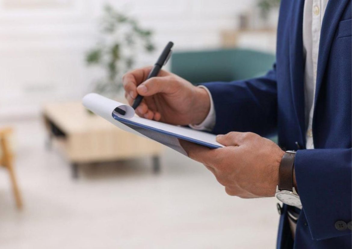 Un agent immobilier en costume bleu prend des notes sur un presse-papiers lors d'une visite.