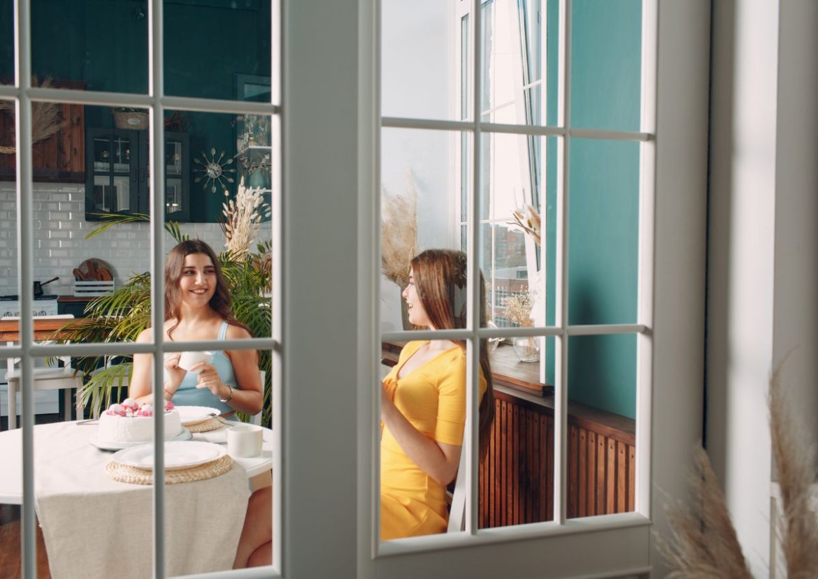 Moment de convivialité entre amies vues derrière des fenêtres lumineuses. Deux femmes souriantes partageant un moment de détente et un dessert, vues à travers des fenêtres à carreaux.