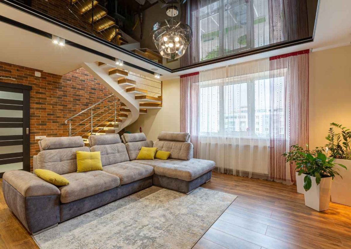 Design intérieur contemporain et efficacité énergétique. Salon moderne et lumineux avec escalier design, canapé d'angle gris et briques murales, reflétant l'importance du confort après une rénovation énergétique.