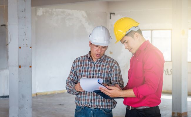 Deux ingénieurs ou chefs de chantier discutant de plans sur une feuille de papier, dans un bâtiment en cours de construction.