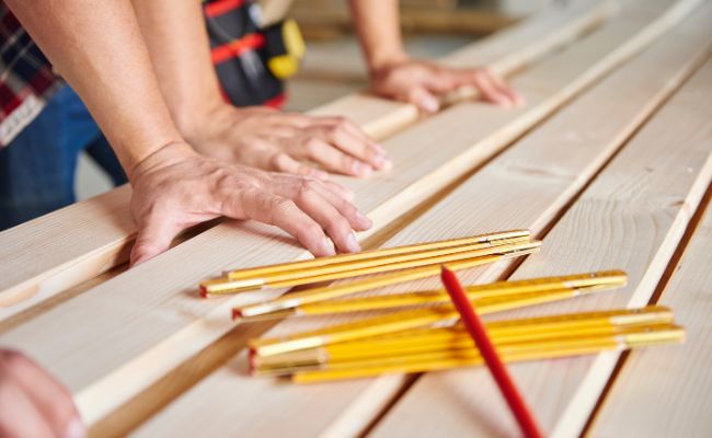 Menuiseries : Mains d'artisans touchant des planches de bois brut avec un mètre pliant.
