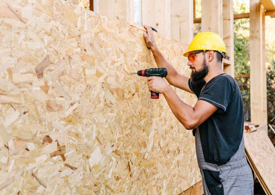 Ouvrier en tenue de travail et casque de sécurité, installant un panneau OSB, première étape de l'isolation extérieure d'une maison en construction.
