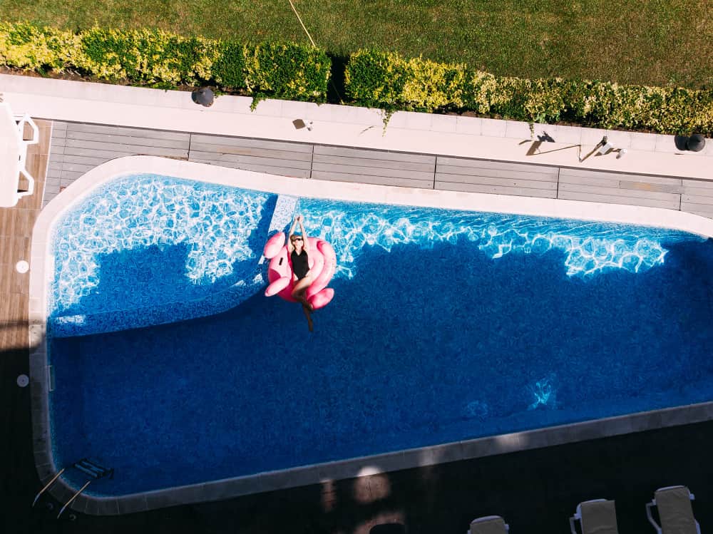 Vue aérienne d'une piscine coque de forme libre avec de l'eau bleue, une personne flottant sur une bouée flamant rose géante dans un jardin ensoleillé
