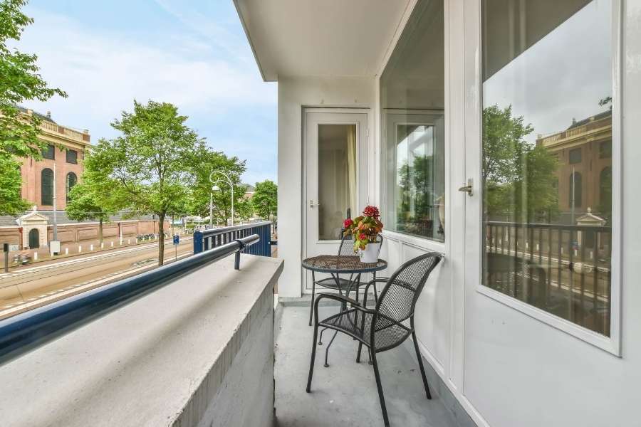 Un petit balcon étroit en ville avec une table et deux chaises en métal noir, donnant sur une rue bordée d'arbres.